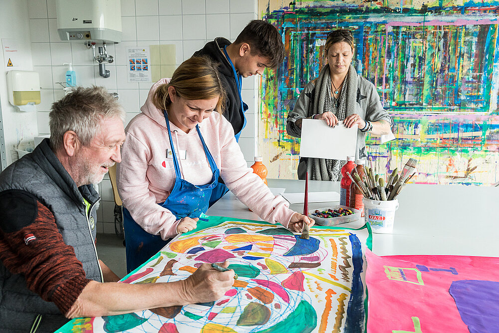 Ein älterer Mann und eine junge Frau mit blauer Schürze arbeiten gemeinsam an einem Bild, das aus vielen bunten Farbflächen besteht. Im Hintergrund sind zwei weitere Personen zu sehen, die ebenfalls künstlerisch tätig sind. 