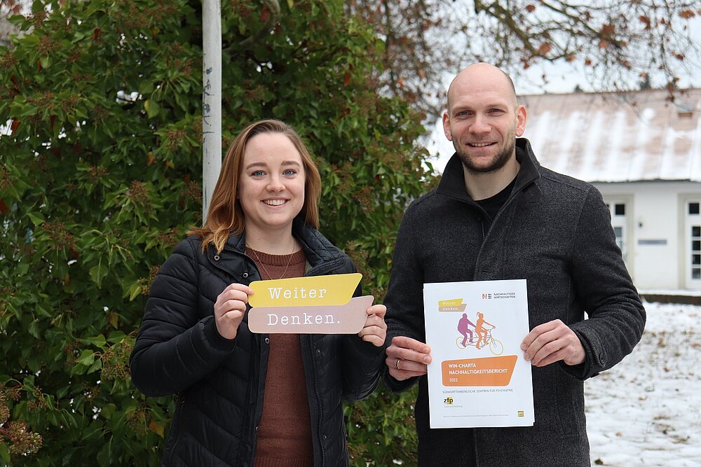 Eine junge Frau und ein junger Mann stehen nebeneinander. Sie hält ein Logo mit dem Text Weiterdenken in der Hand, er ein Din-A-4 Dokument, auf dem Nachhaltigkeitsbericht zu lesen ist. 