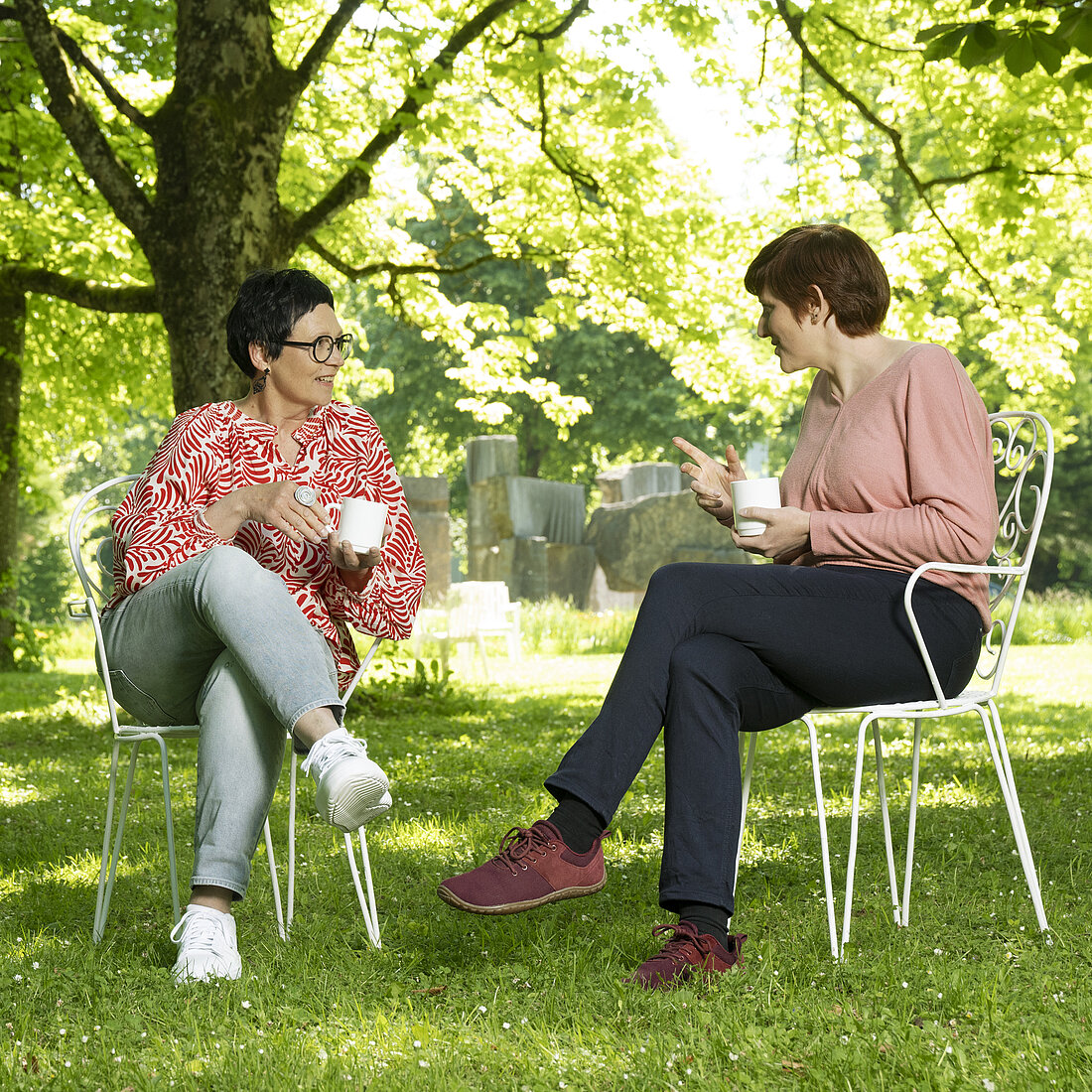 Zwei Frauen sitzen auf weißen Metallstühlen in einem grünen Park und trinken aus weißen Tassen.