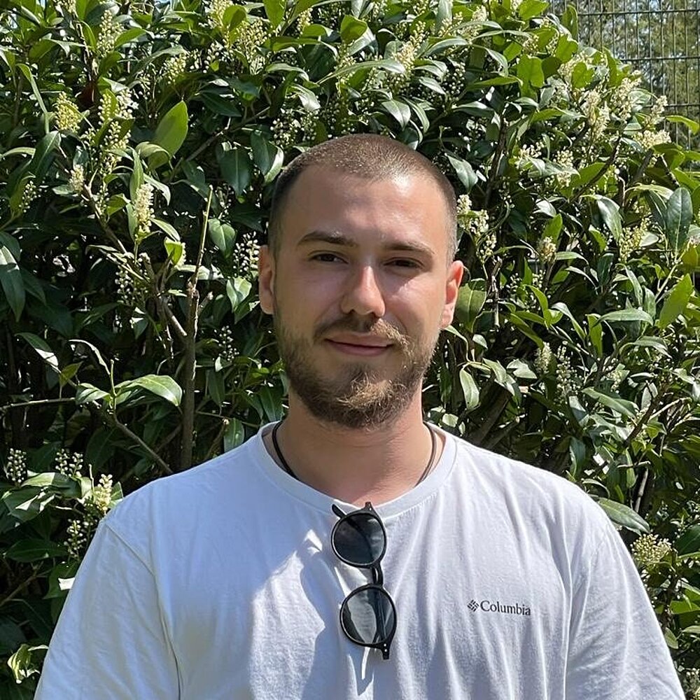 Ein junger Mann steht mit weißem T-Shirt und eingehakter Sonnenbrille vor einer Hecke.