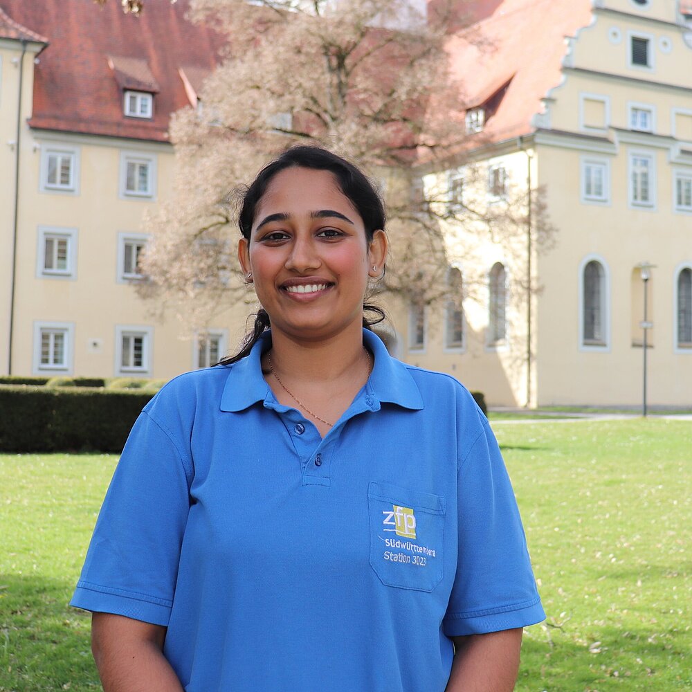 Eine junge Inderin in Arbeitskleidung des ZfP steht vor dem Münster in Zwiefalten und schaut lächelnd in die Kamera. 