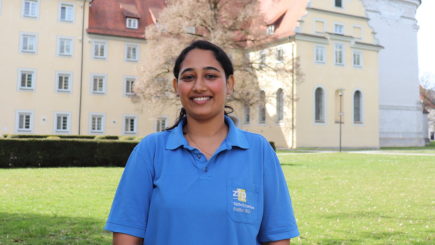 Eine junge Inderin in Arbeitskleidung des ZfP steht vor dem Münster in Zwiefalten und schaut lächelnd in die Kamera. 