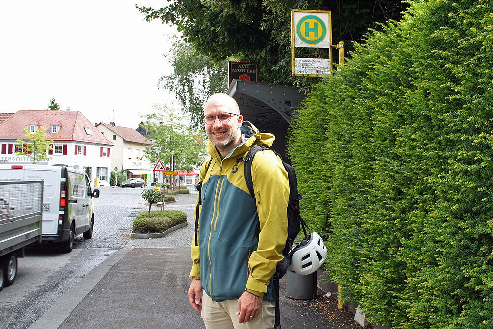 Ein Mann mit Brille, bepackt mit Rucksack und Fahrradhelm, wartet an einer Bushaltestelle.