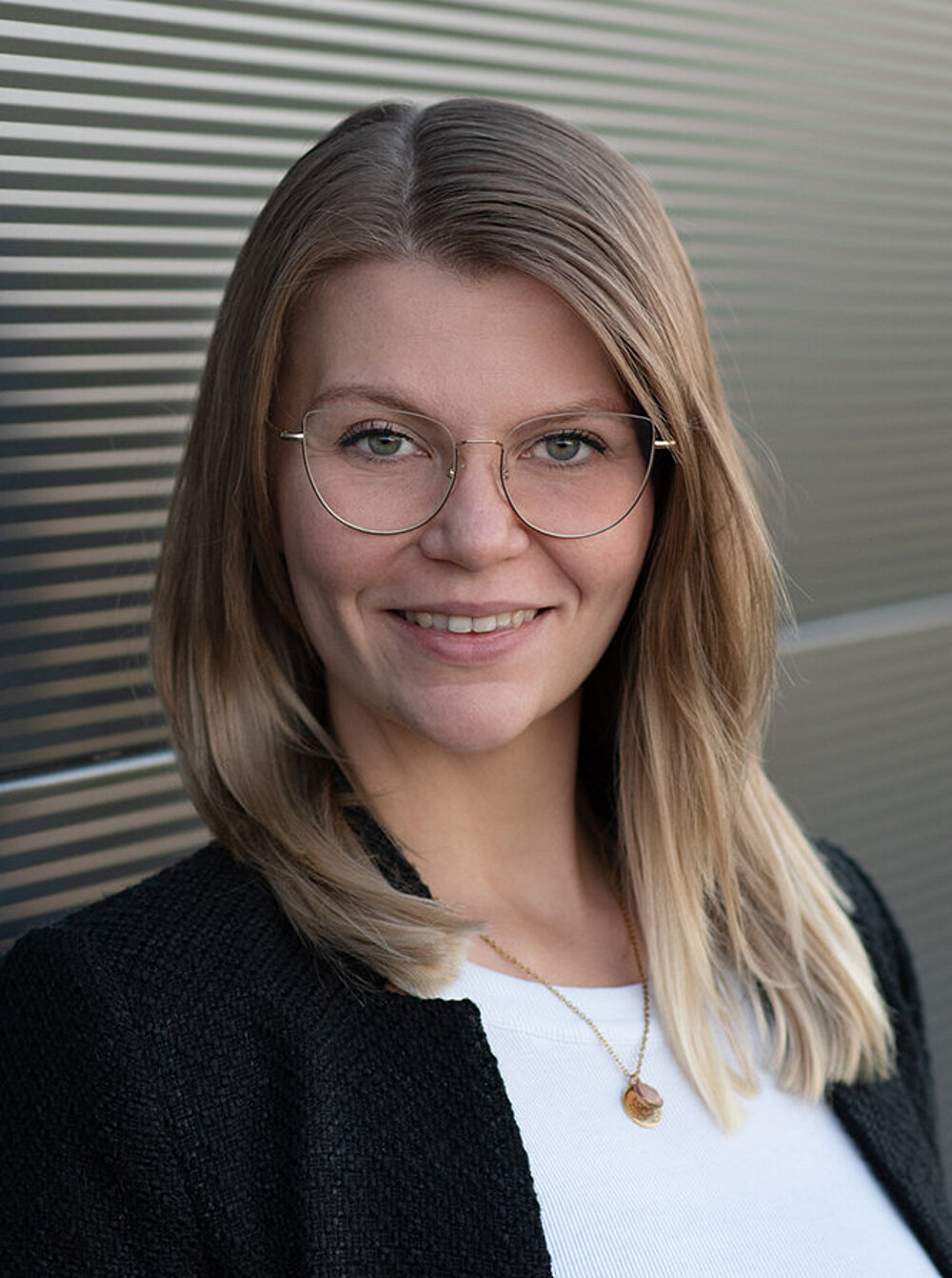 Eine Frau mit blonden langen Haaren, Brille und weißer Bluse schaut lächelnd in die Kamera.