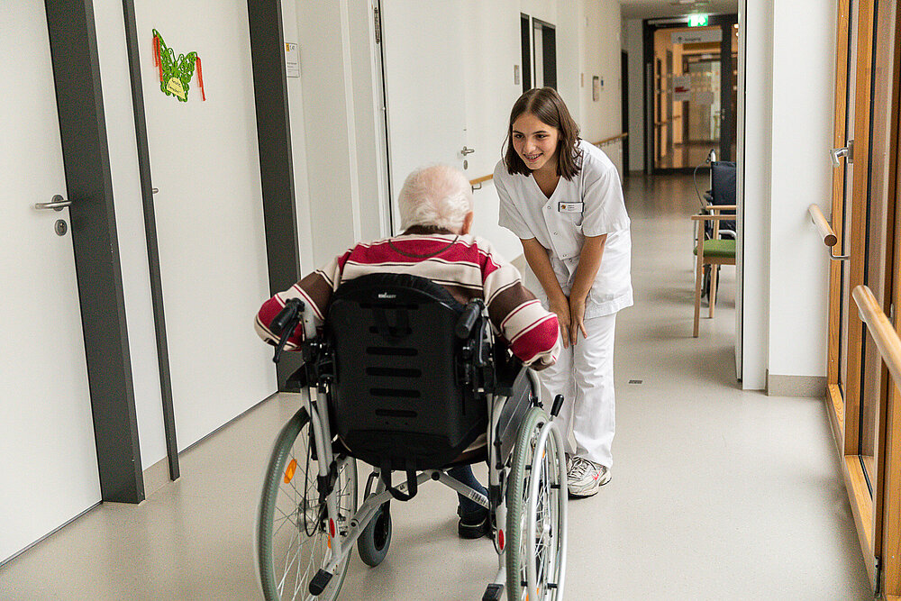 Eine junge Pflegekraft beugt sich lächelnd zu einem älteren Mann im Rollstuhl hinunter.
