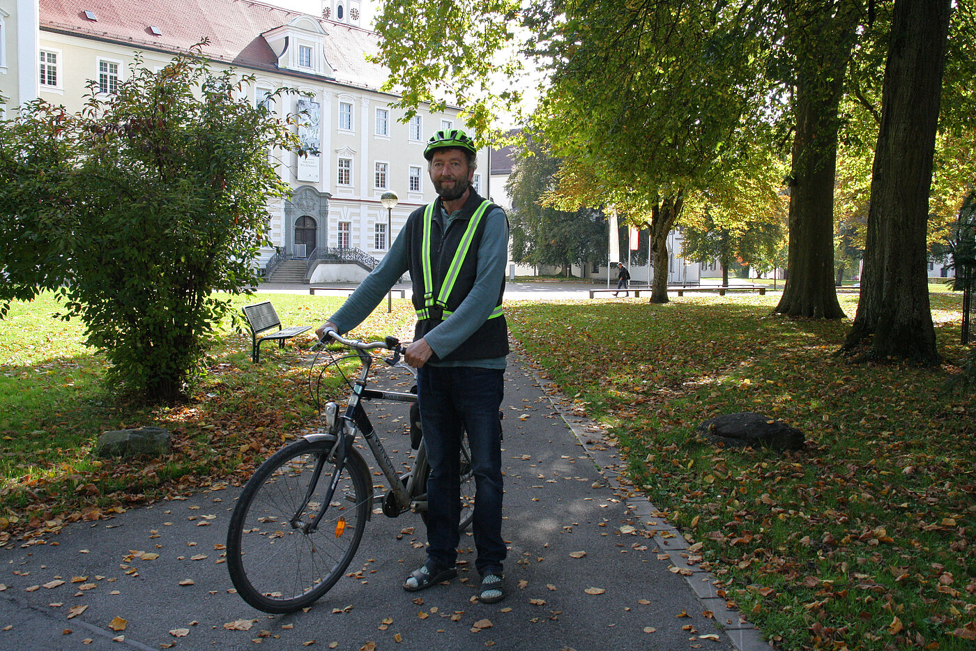 Ein Mann mittleren Alters mit Helm und Reflektorweste steht auf einem von Bäumen gesäumten Weg und hält mit den Händen sein Fahrrad.
