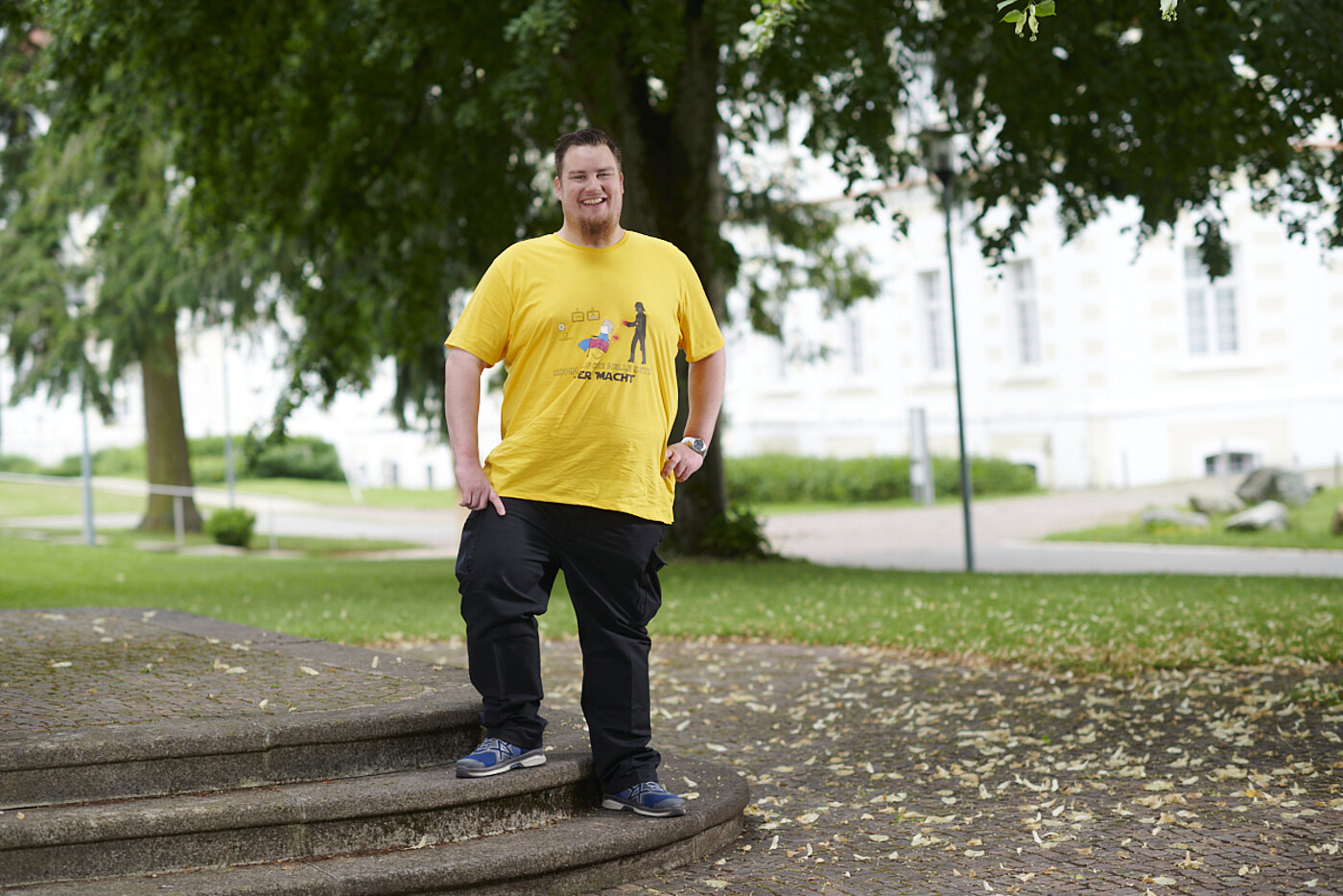 Ein kräftiger Mann mit gelbem T-Shirt steht lachend auf einer kleinen Treppe im Freien und stemmt die Arme in die Hüften.