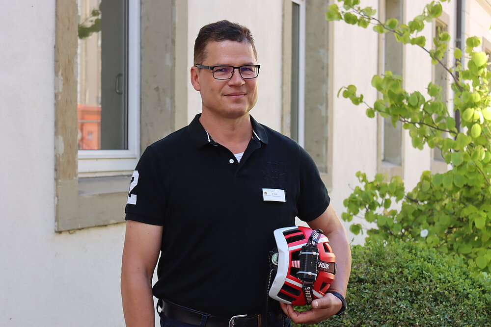 Ein junger Mann mit kurzen, dunklen Haaren und Brille, der einen Fahrradhelm unter dem Arm hat. 