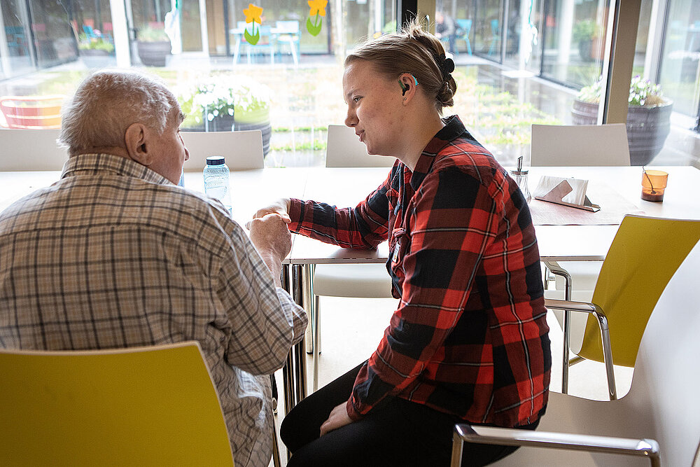 Eine junge Pflegekraft hält sitzt mit einem Patienten am Tisch und hält seine Hand. 