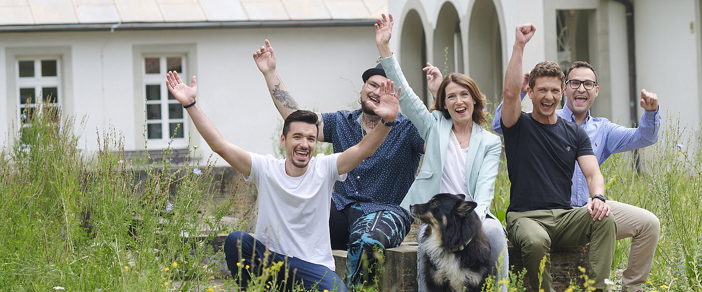 Eine Frau, vier Männer und ein Hund sitzen nebeneinander und recken fröhlich die Arme in die Höhe. Um sie herum ist eine grüne Naturwiese zu sehen, im Hintergrund ein weißes Gebäude. 