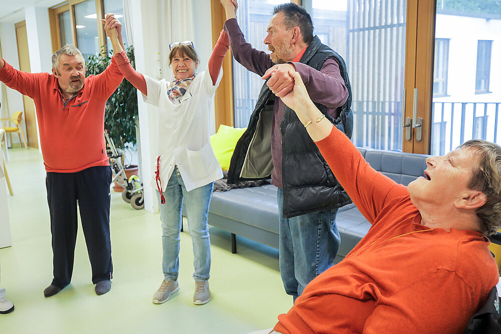 Eine Frau mit Berufskleidung bildet mit mehreren älteren Menschen eine Kette. Die Gruppe hebt die ineinandergelegten Hände in die Höhe. Die Pflegerin lacht fröhlich. 