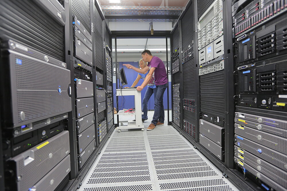 Man blickt in einen großen Serverraum, an dessen Wänden sich zahlreiche Rechner türmen. Im Hintergrund stehen eine Frau und ein Mann und blicken auf einen Monitor. 