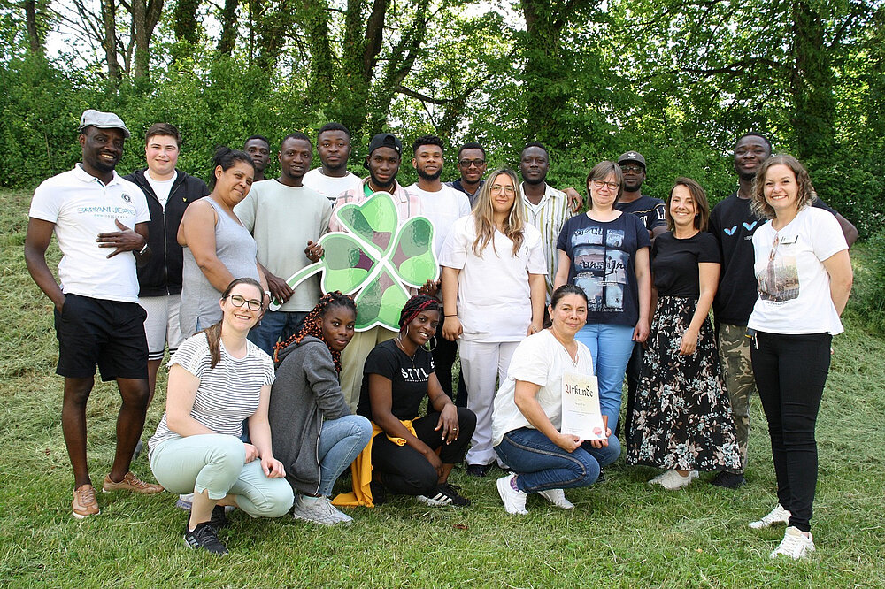 Auf einer Wiese hat sich eine Gruppe junger Menschen für ein Gruppenfoto aufgestellt. Manche von ihnen knien vorne im Gras. Hinter der Gruppe stehen Laubbäume.