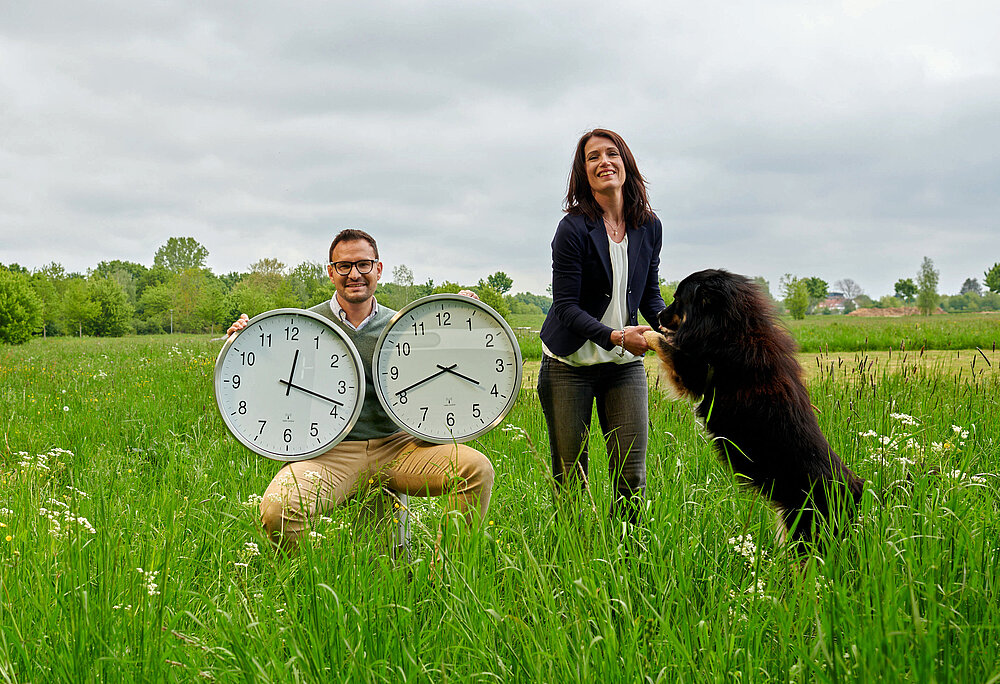 Ein junger Mann hält zwei große Uhren unterm Arm, eine junge Frau spielt im Vordergrund mit ihrem Hund. 