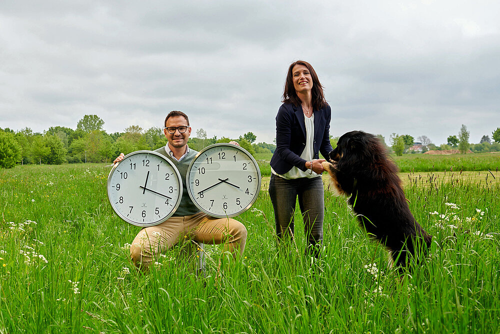 Ein junger Mann hält zwei große Uhren unterm Arm, eine junge Frau spielt im Vordergrund mit ihrem Hund. 