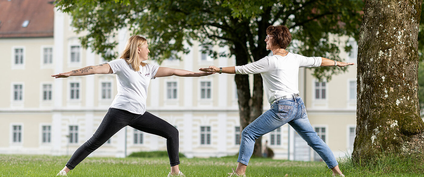 Eine jüngere und eine ältere Frau stehen auf einer grünen Wiese. Sie stehen sich in der Yogahaltung Krieger gegenüber. 