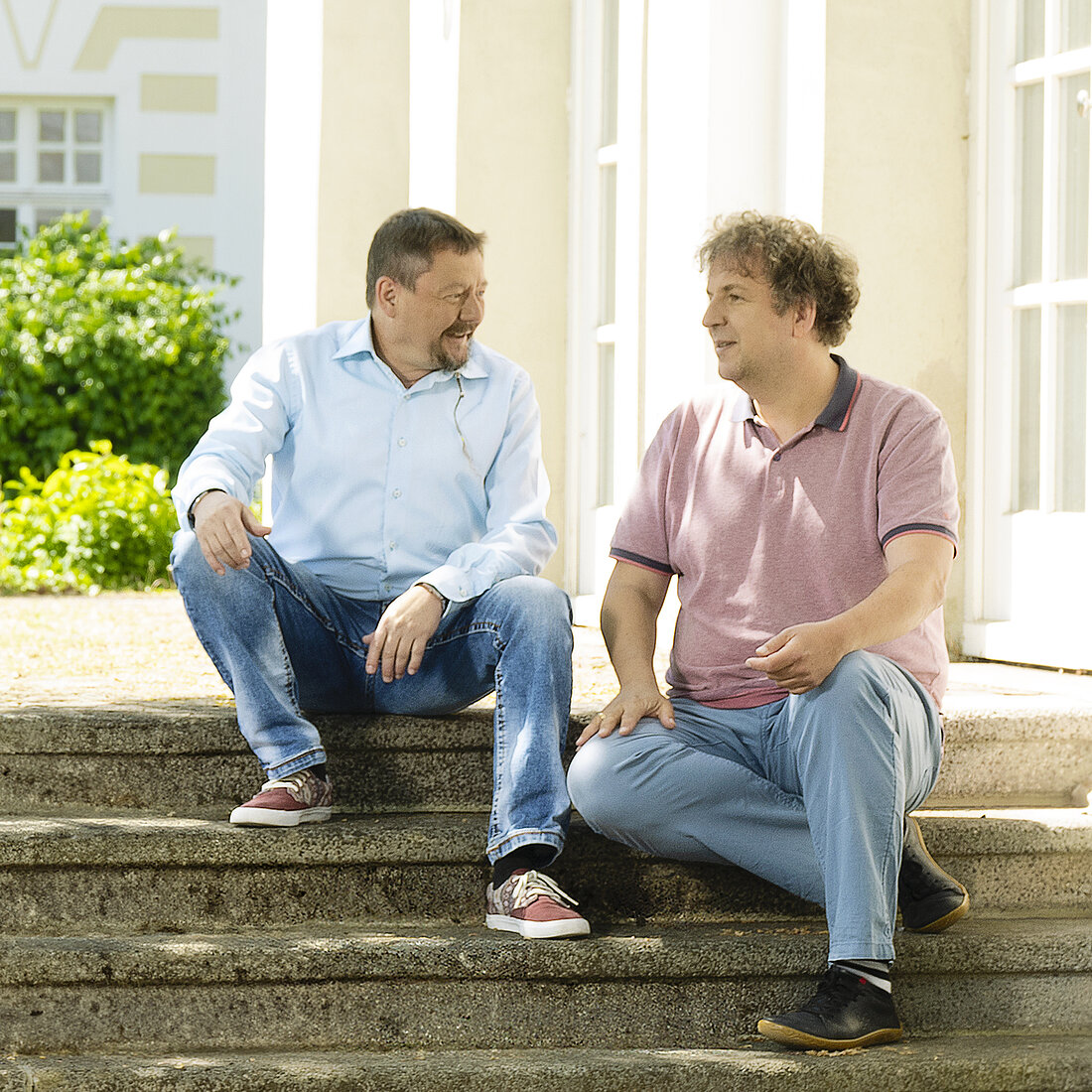 Zwei Männer sitzen auf Außentreppen vor einem hellen Gebäude, einer trägt ein hellblaues Hemd und Jeans, der andere ein rosa Poloshirt und hellblaue Hose.