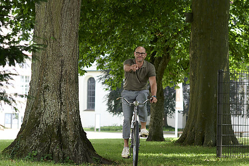Ein Mann fährt auf dem Fahrrad einen von Bäumen umsäumten Weg entlang, lacht und zeigt mit der rechten Hand in die Kamera.