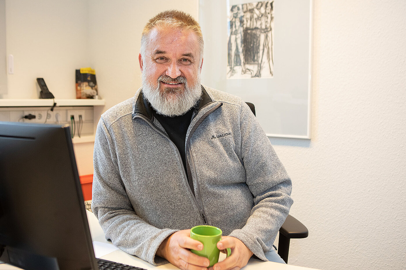 Ein älterer Mitarbeiter der Pflege mit kurzen grauen Haaren und Vollbart sitzt lächelnd mit Kaffeetasse in der Hand an einem Schreibtisch.