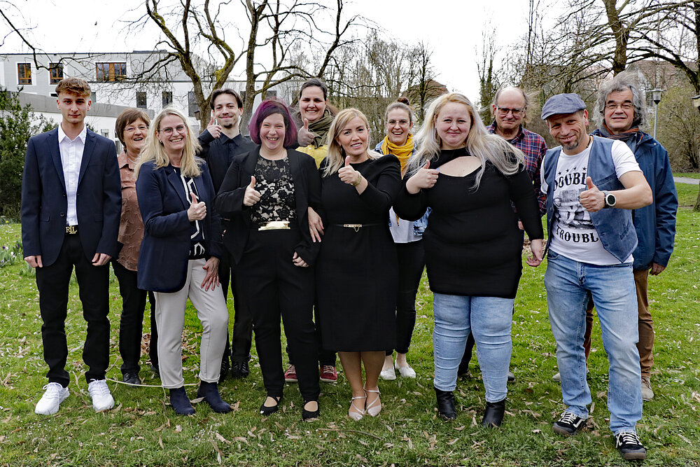 Eine Gruppe junger Menschen, teilweise in festlicher Kleidung, steht auf einer grünen Wiese. 