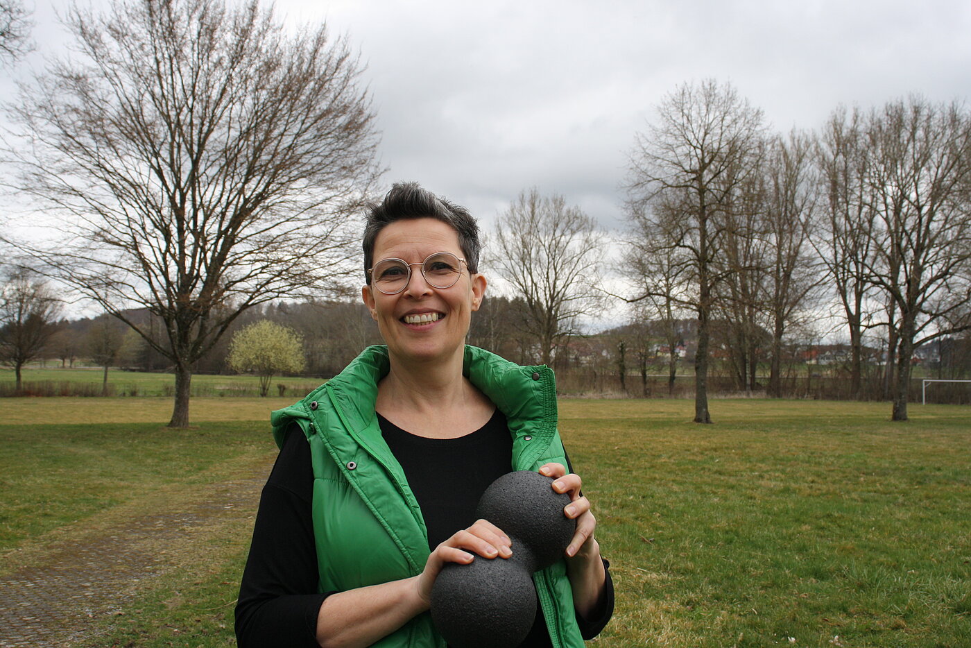 Eine Frau mit Brille und kurzen schwarzen Haaren hält auf einer Wiese stehend lachend eine Faszienrolle in den Händen.