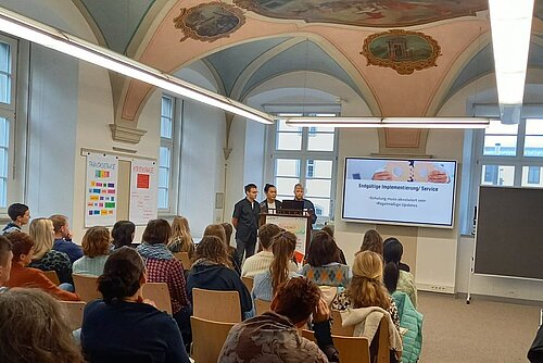 In einem historischen Klostergebäude referieren drei junge Männer vor Publikum. Sie stehen neben einer Beamer-Leinwand.