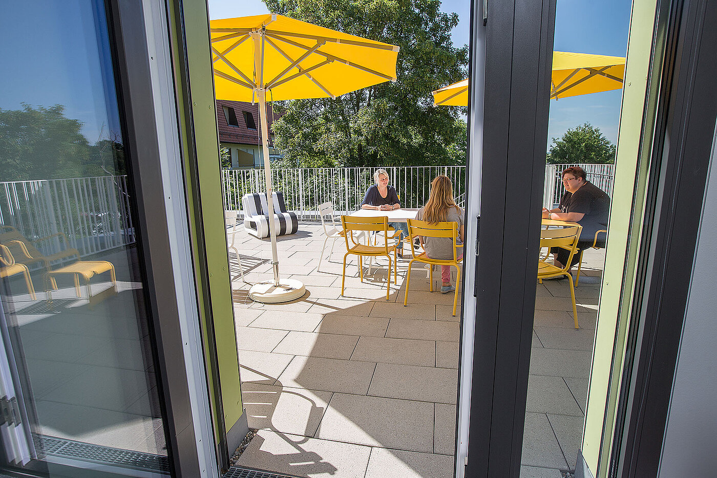 Durch eine geöffnete Terassentür sieht man auf einen Balkon mit gelben Sonnenschirmen. 