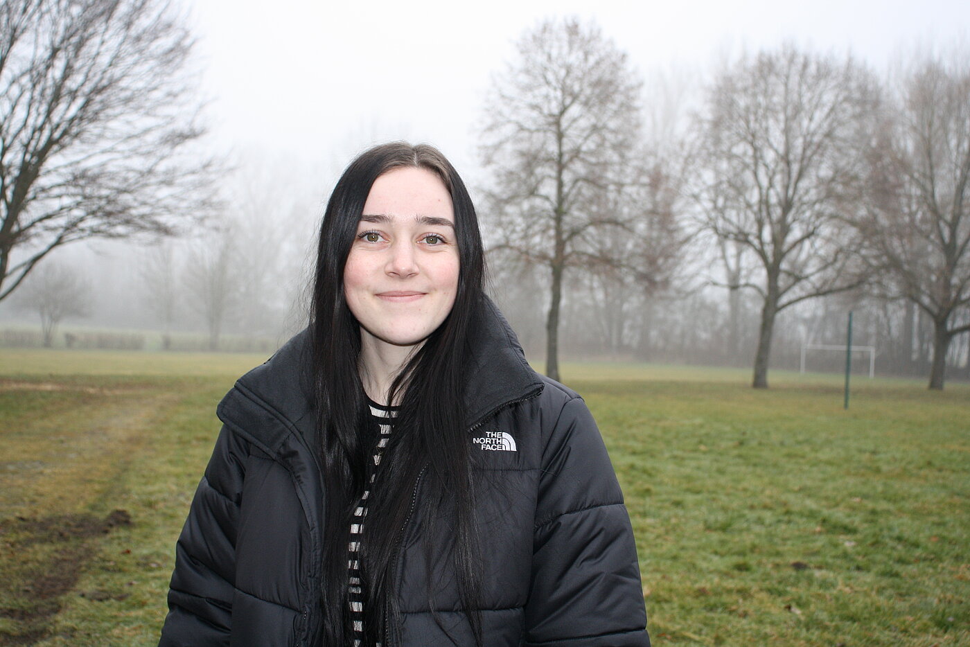 Eine junge Frau mit langen schwarzen Haaren und schwarzer Winterjacke steht lächelnd auf einer nebligen Wiese.