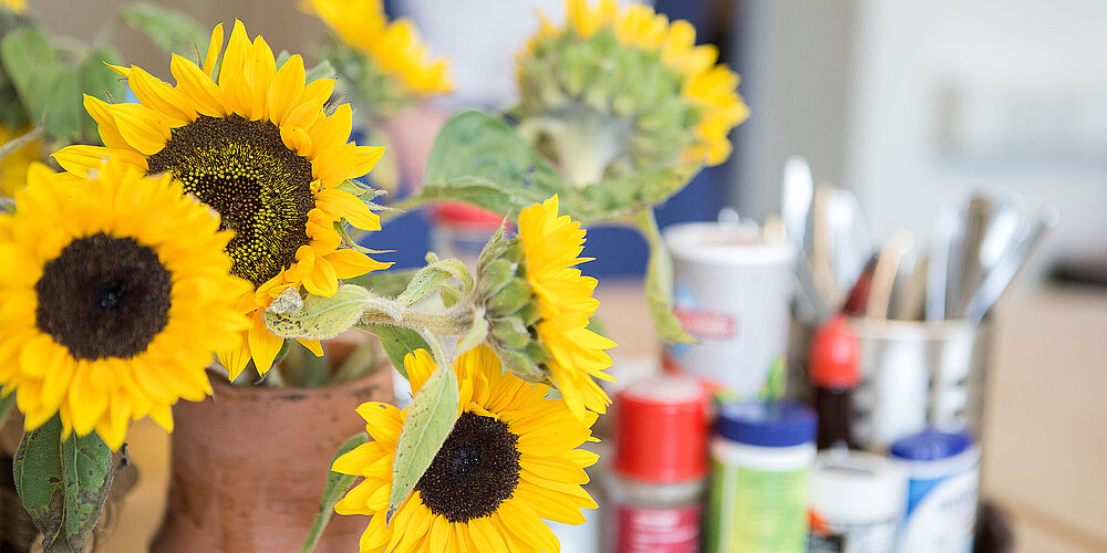 Im Vordergrund steht ein Strauß mit großen Sonnenblumen, im Hintergrund ein Tablett mit Malutensilien. 