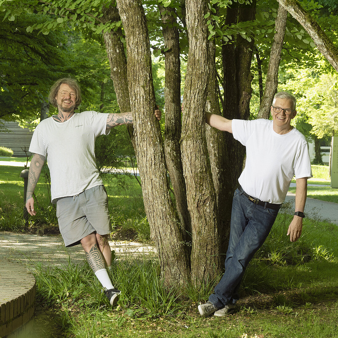 Zwei Männer lehnen entspannt an einem Baum in einem Park mit grünem Gras und Bäumen.