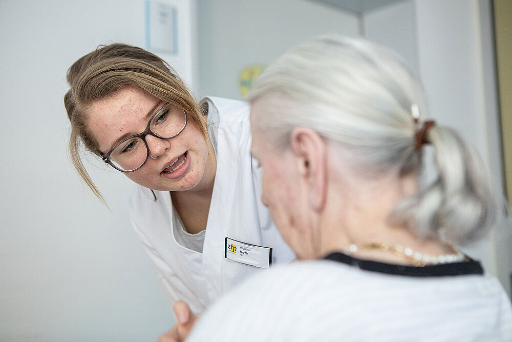 Eine junge Frau mit Brille beugt sich zu einer alten Frau hinunter.