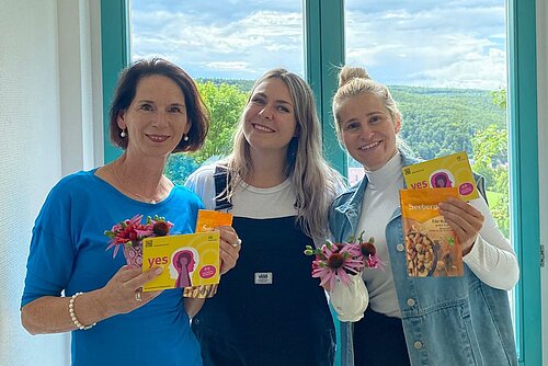 Drei Frauen stehen nebeneinander, schauen lachend in die Kamera und halten Werbematerial zum Podcast des ZfP Südwürttemberg in den Händen.