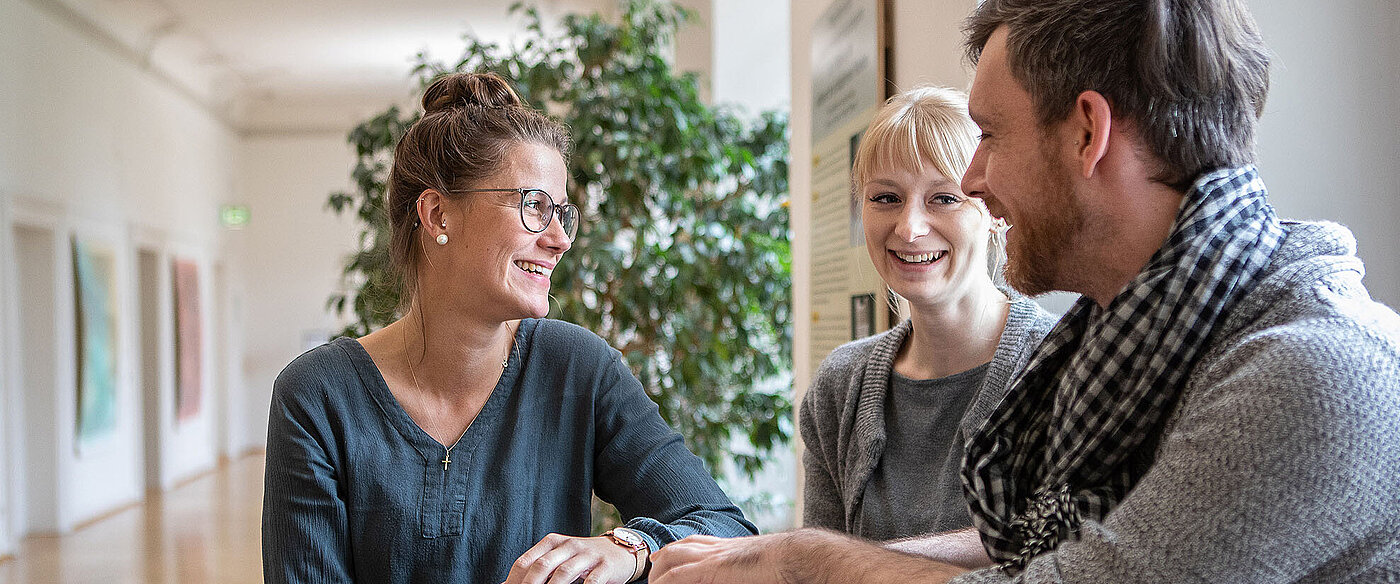 Zwei Frauen und ein Mann sitzen an einem Bistrotisch, auf dem Seminarunterlagen liegen. 