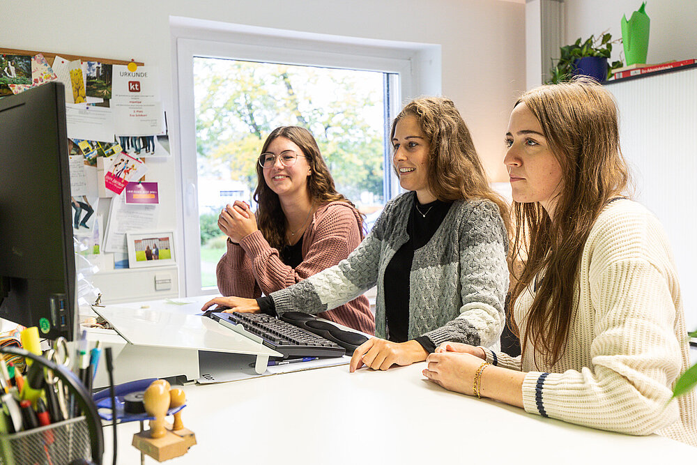 Drei Frauen stehen an einem Schreibtisch und schauen gemeinsam in einen Bildschirm.