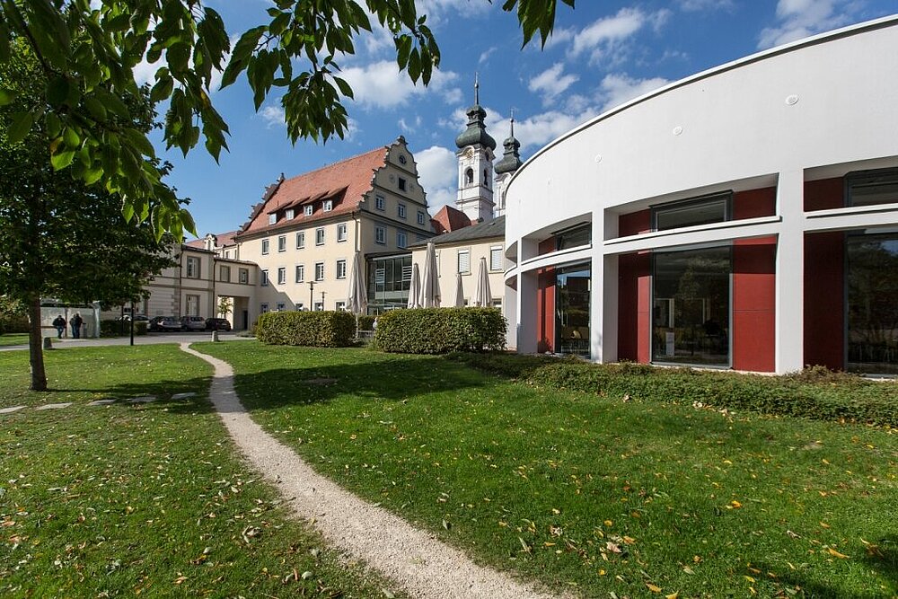 Ein Klostergebäude und modernes rundgeschnittenes Gebäude sind im Hintergrund, im Vordergrund ein Weg und eine Wiese zu sehen.