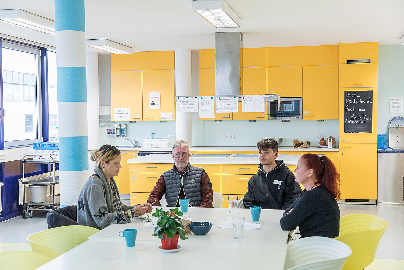 Vier Menschen sitzen an einem Tisch, im Hintergrund ist eine offene Küche zu sehen. 