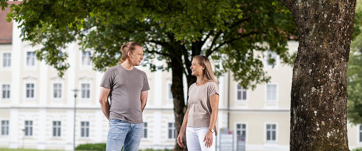 Eine Gruppe Menschen steht vor einem Baum und begrüßt eine junge Frau, die auf einem E-Scooter steht und lacht. 