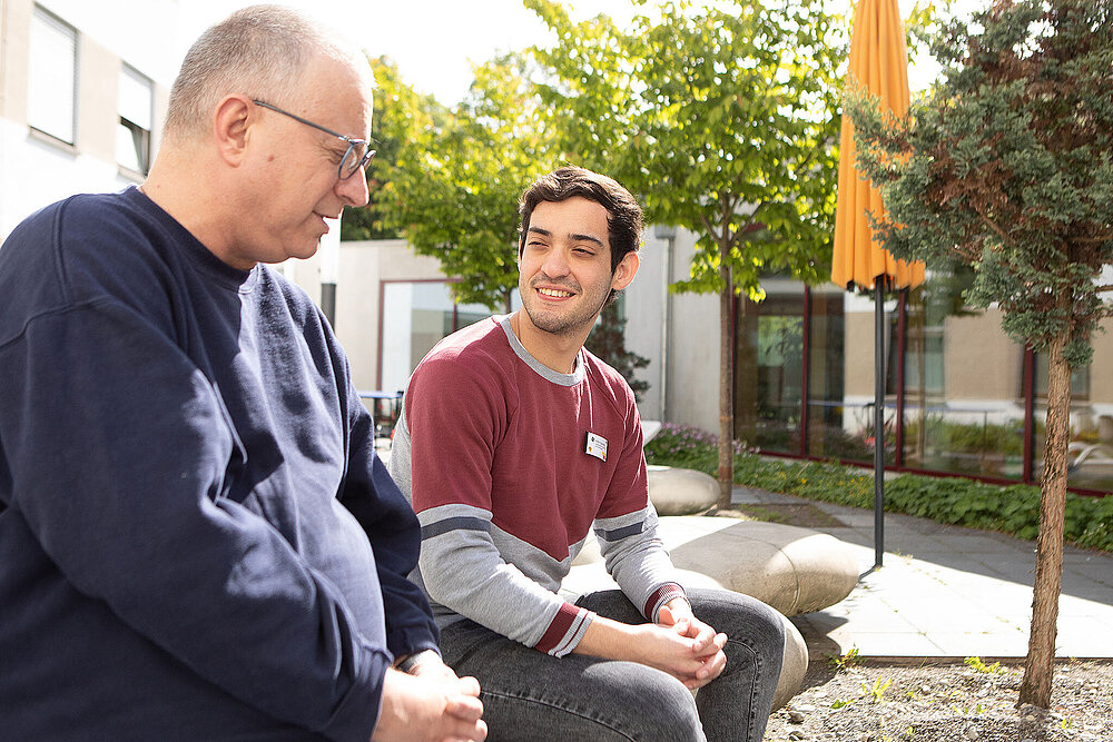 Ein jungen Mann sitzt mit einem Patienten auf einer Bank und schaut ihn von der Seite an.