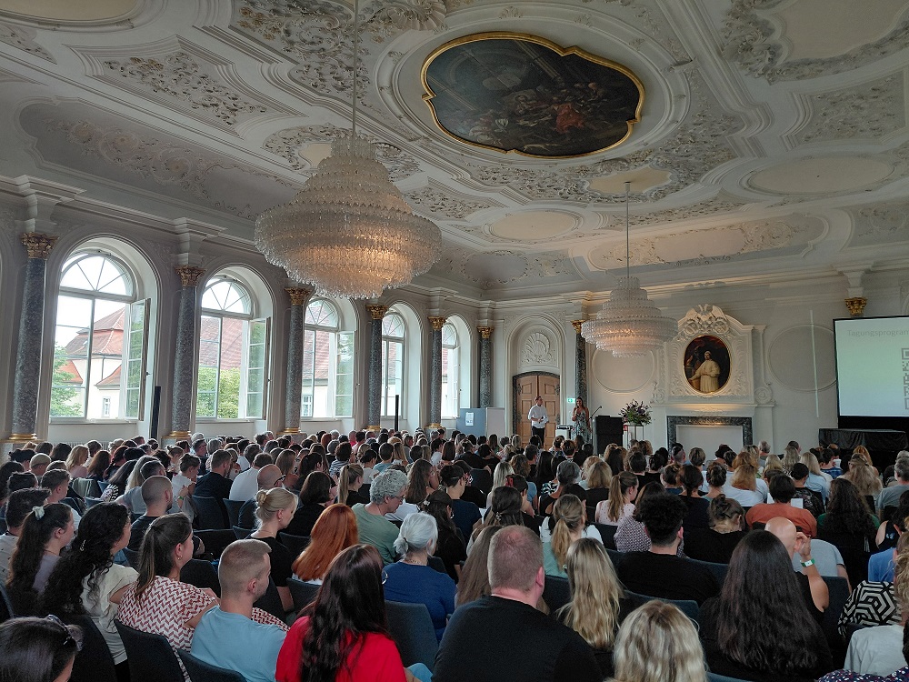 Ein vollbesetzter und bestuhlter Klostersaal, die Teilnehmenden sind von hinten und seitlich zu sehen und blicken auf eine kleine Bühne nach vorne, wo zwei Personen moderieren.